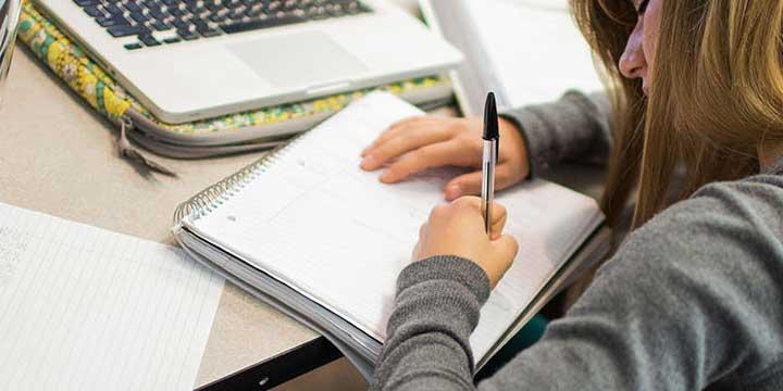 Young woman writing in spiral notebook on desk infront of laptop